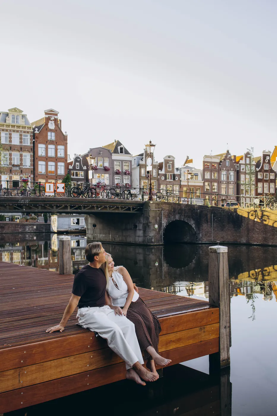 Couple on a canal pontoon in Amsterdam with canal houses and a bridge behind