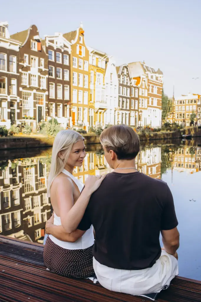 Couple sitting on a wooden pontoon on an Amsterdam canal, gabled canal houses reflected in the water