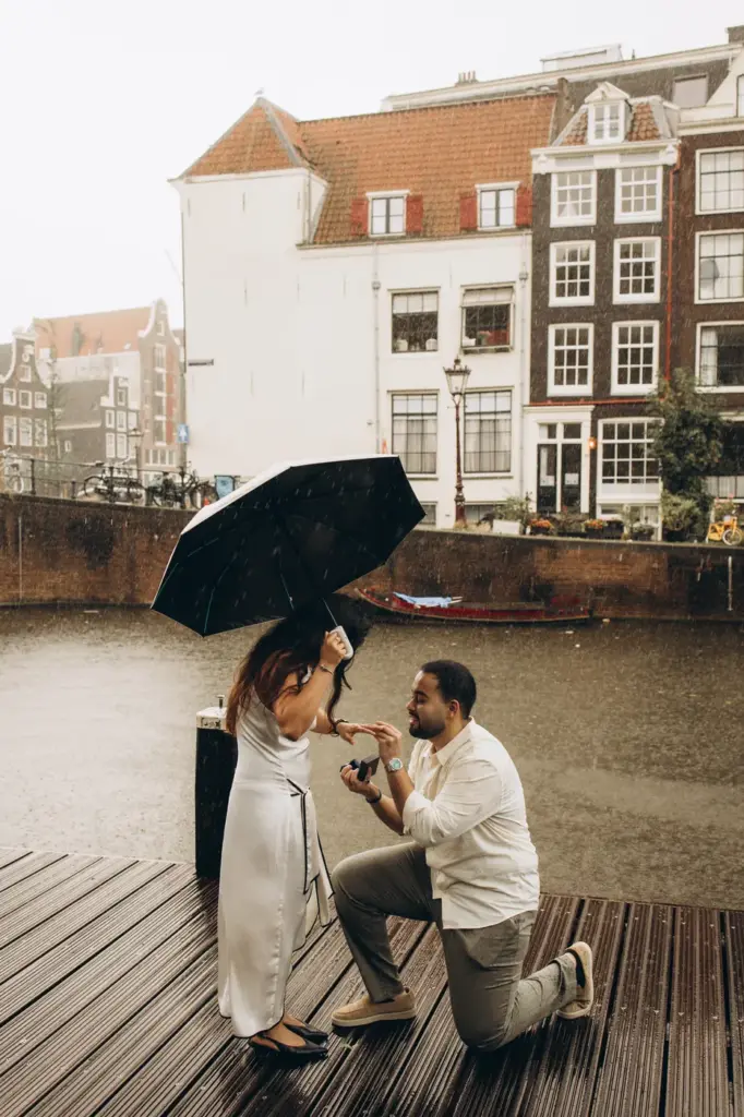Man kneeling on a rainy canal pontoon in Amsterdam, proposing under an umbrella